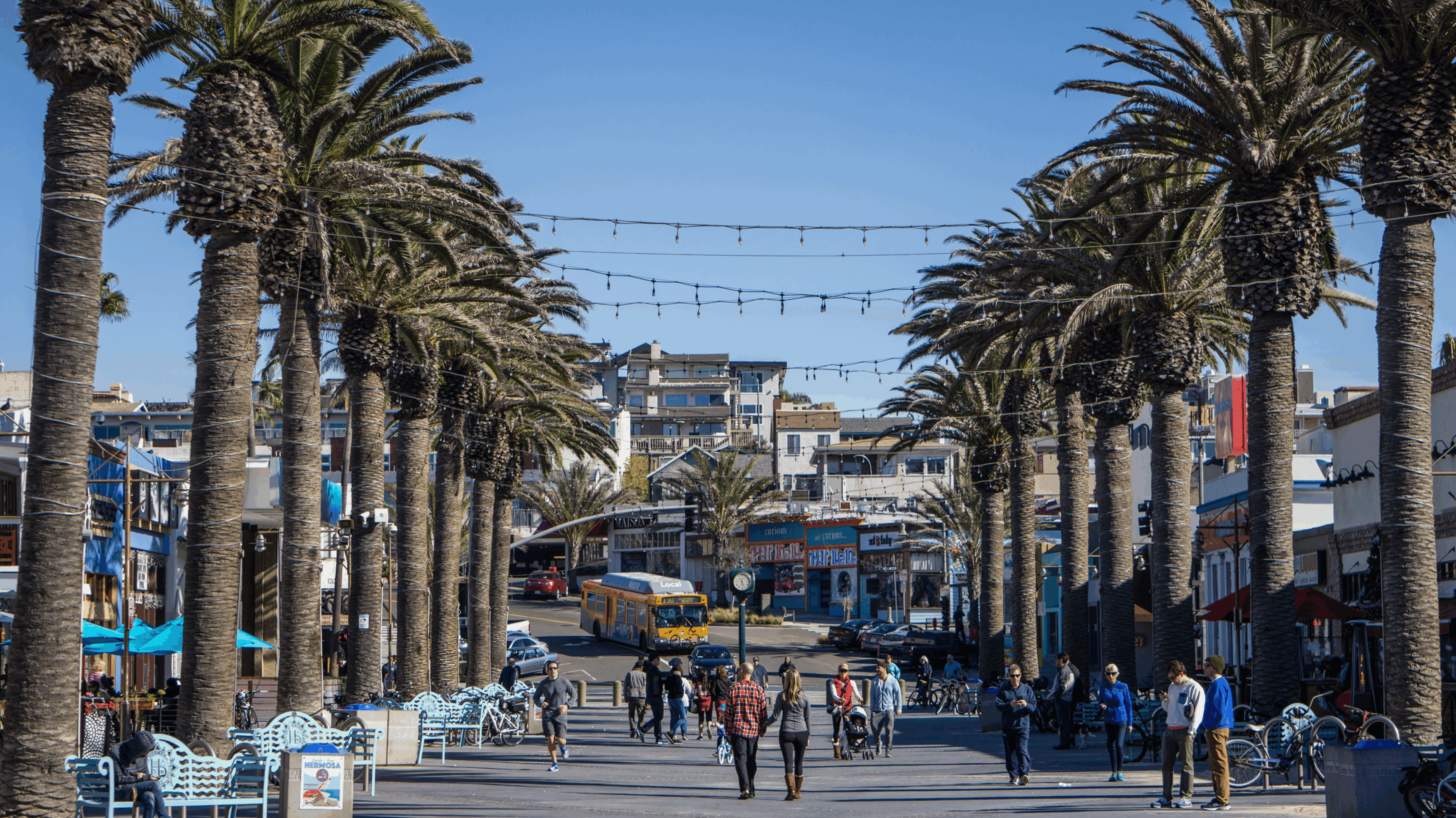 People walking along a palm-lined street with buildings and string lights overhead.