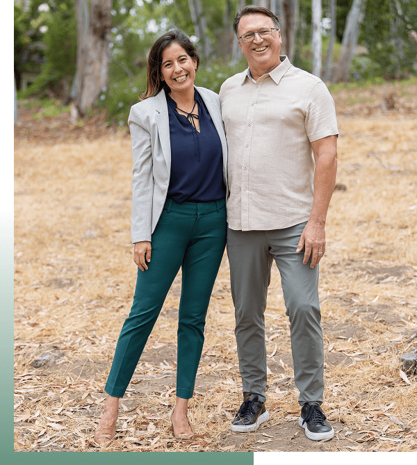 A smiling couple stands together outdoors on a dirt path.